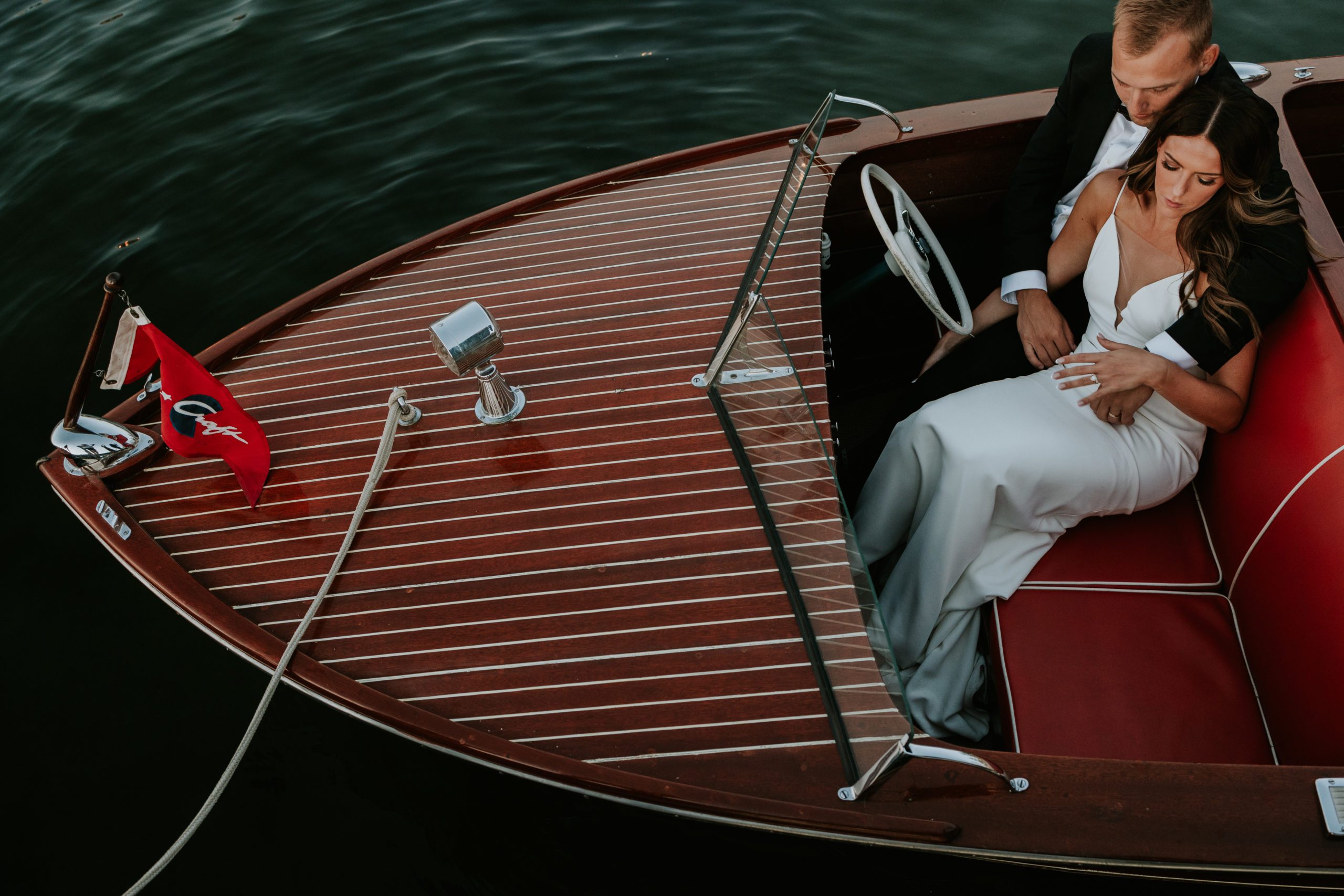 Wedding couple on wooden speedboat in dark shiny waters in lake Okoboji. They are snuggling up on the red bench seat near the steering wheel.