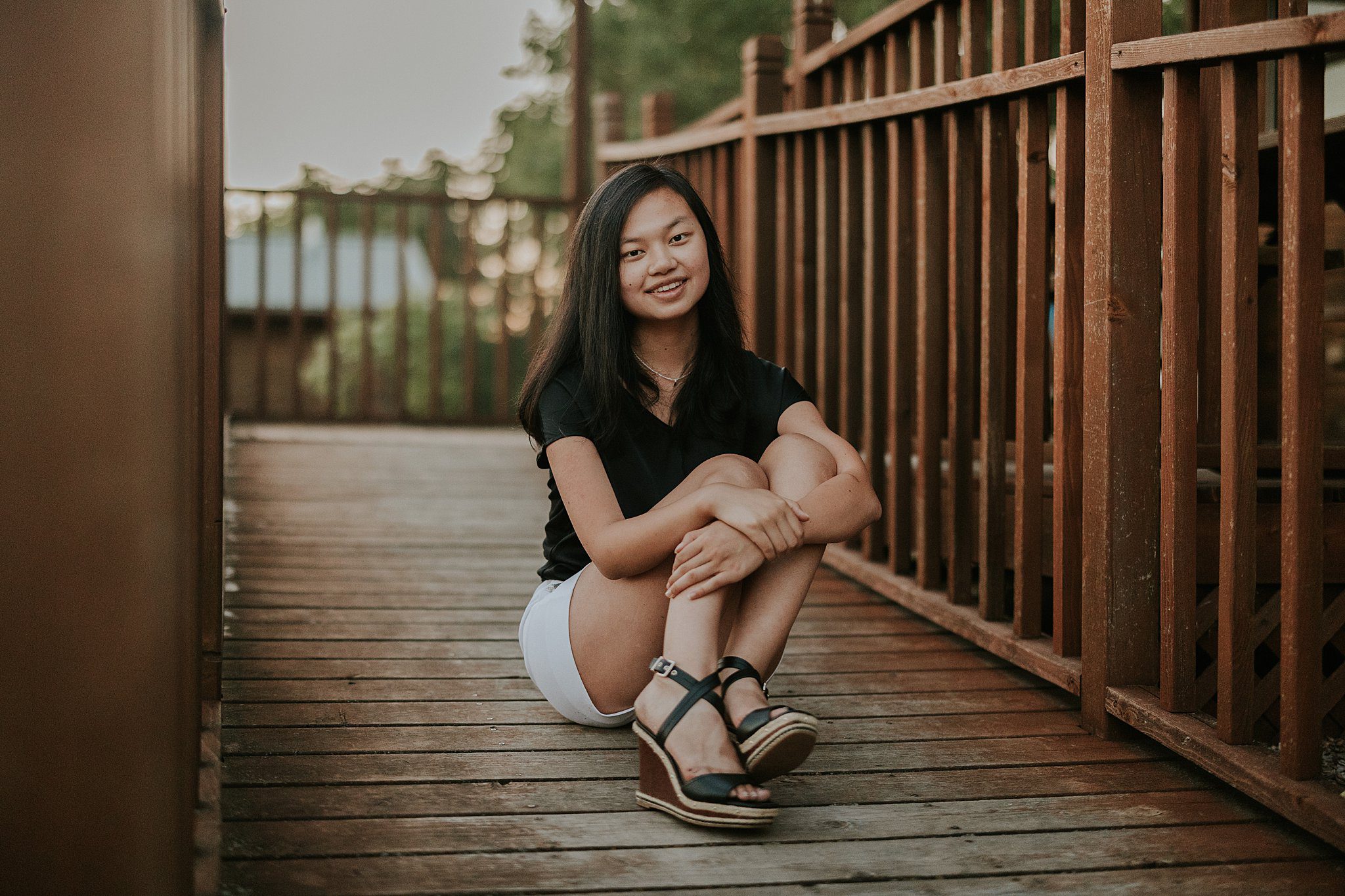 This is a senior portrait taken in front of a cabin in the woods at Oak Grove State Park in Iowa. She is sitting on a wooden ramp  smiling at the camera between stained wooden railings and hugging her knees slightly tilting her knees to the side. She has black hair, a black t shirt, white shorts, and black strappy wedges with one of her feet tipped slightly backwards. 