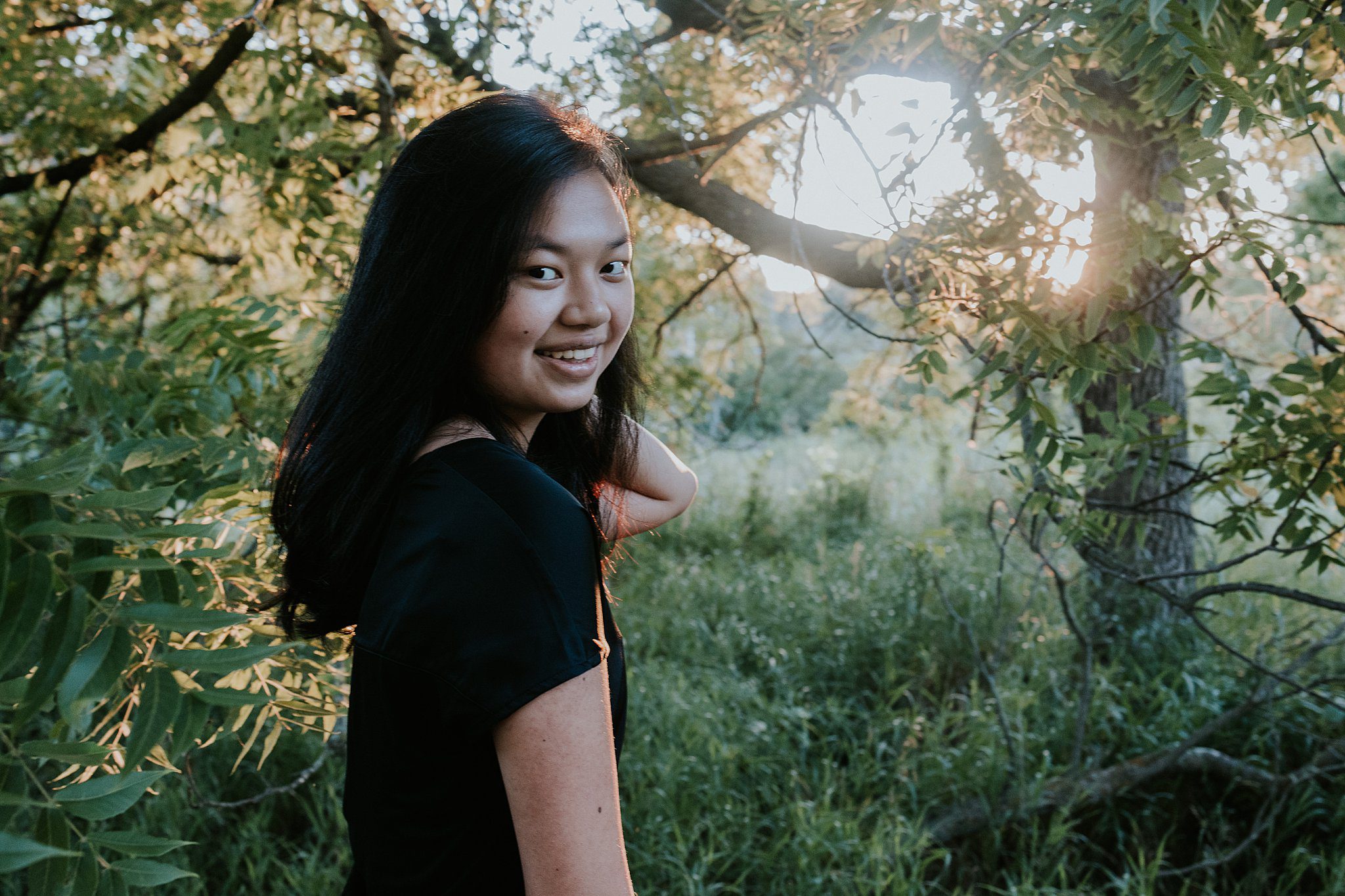 Senior photo of a girl in Northwest Iowa. She is smiling at the camera and her back arm is playing with her hair with her right arm in front of her. The sun is shining through the trees behind her and casting a golden glow.