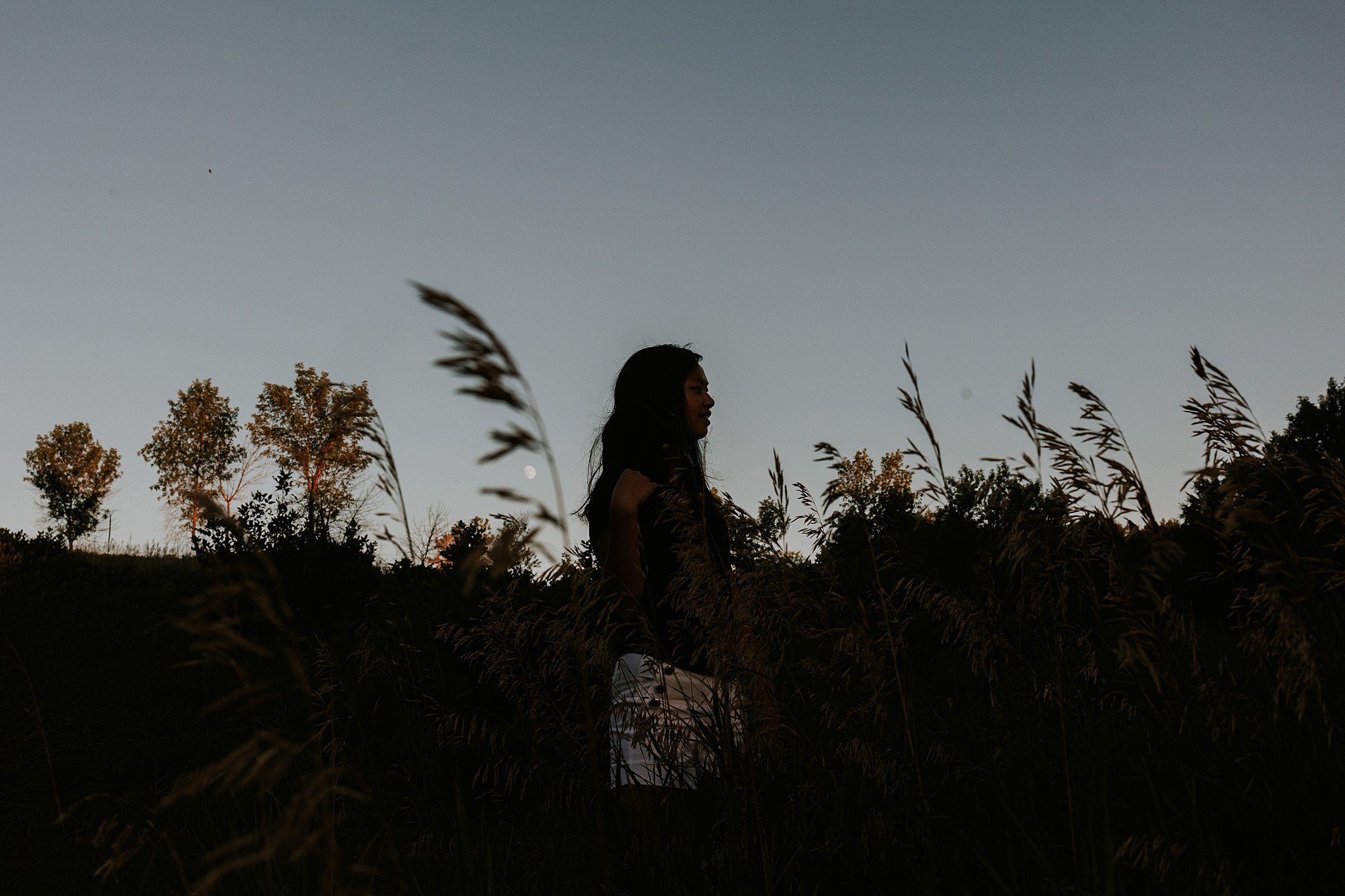 Not your average senior photos. This is a silhouette of a girl looking to the right. The blue sky goes ⅔ down the photo and Wilde grass and side oats fill the bottom of the frame. You can see the moon peaking through the grass next to her. 