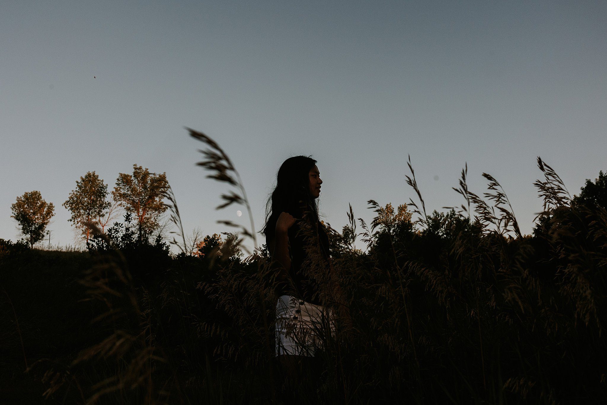 2022-08-09_0005.jpg Not your average senior photos. This is a silhouette of a girl looking to the right. The blue sky goes ⅔ down the photo and Wilde grass and side oats fill the bottom of the frame. You can see the moon peaking through the grass next to her.