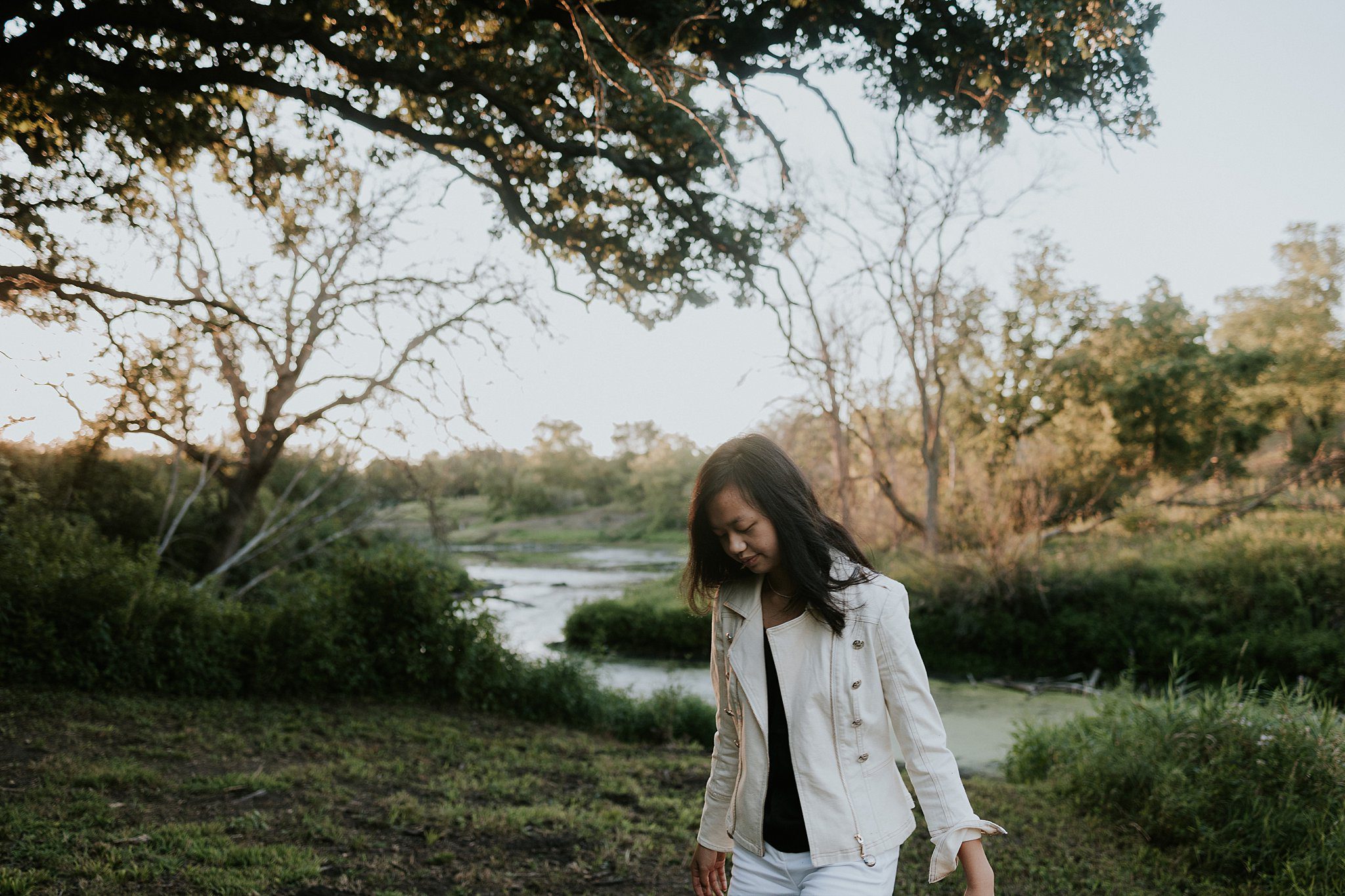 A girl with black hair and a white jacket is walking in front of a pond looking at the ground. 