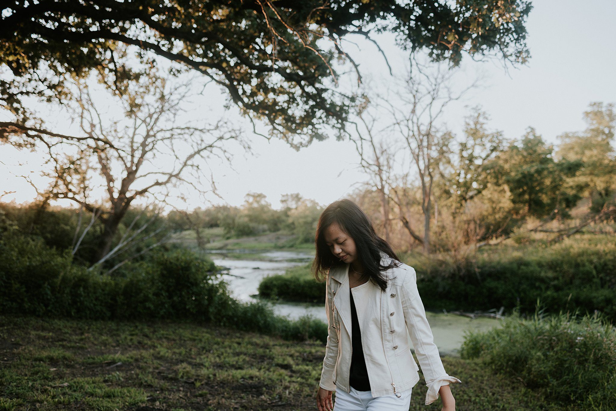 2022-08-09_0007.jpg A girl with black hair and a white jacket is walking in front of a pond looking at the ground.
