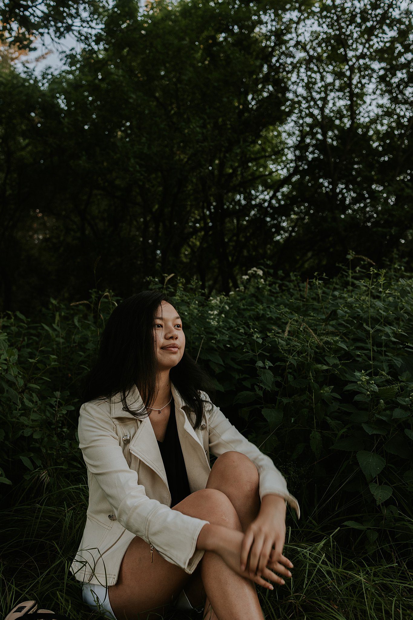 A different senior photo with a girl sitting in the woods. She is looking off to the right and wearing a white leather jacket. 