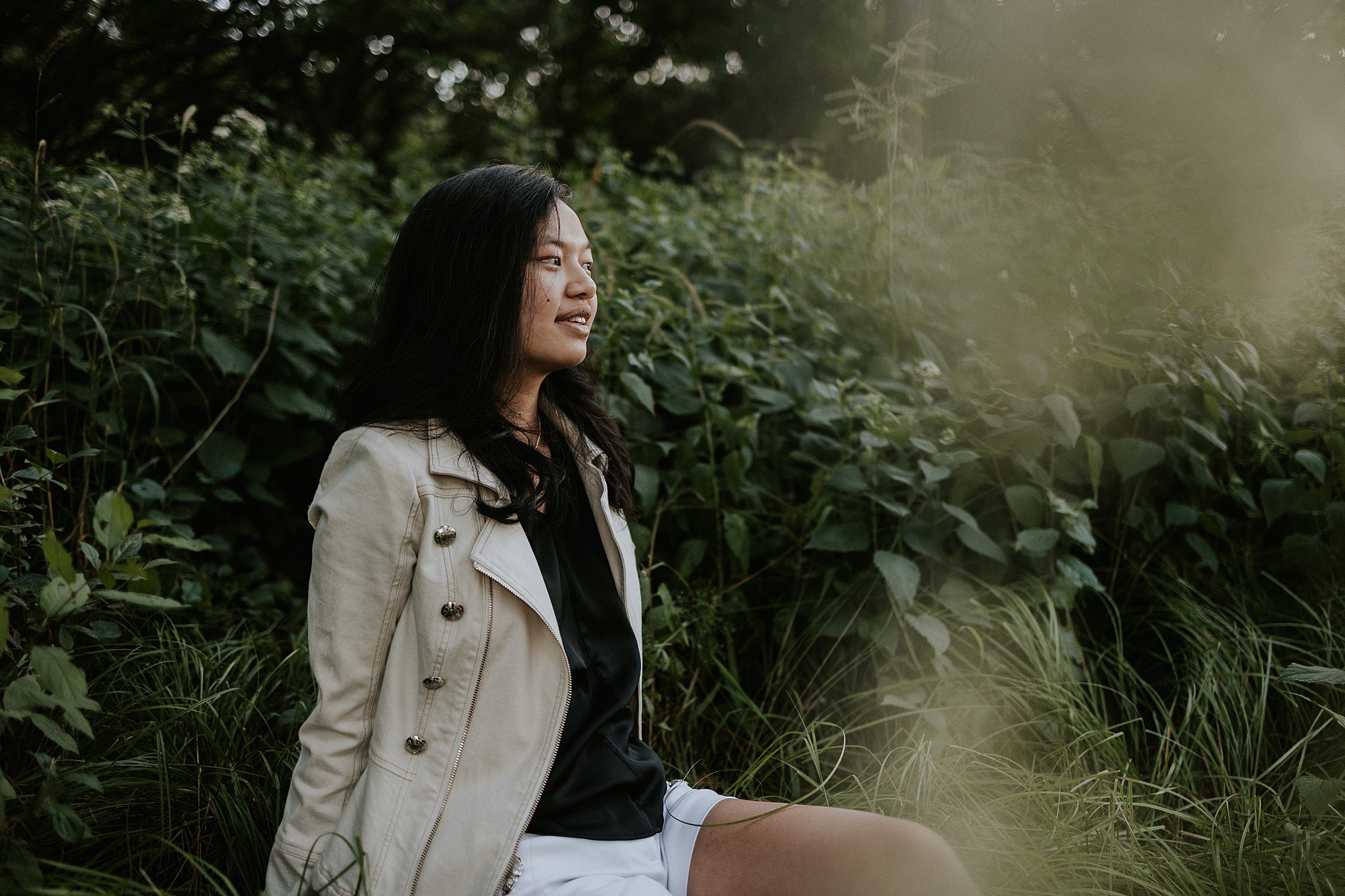 Not boring senior photo. A girl with black hair is sitting leaning back on her arms looking off to the right. There is a layer of plants in front and behind her. 
