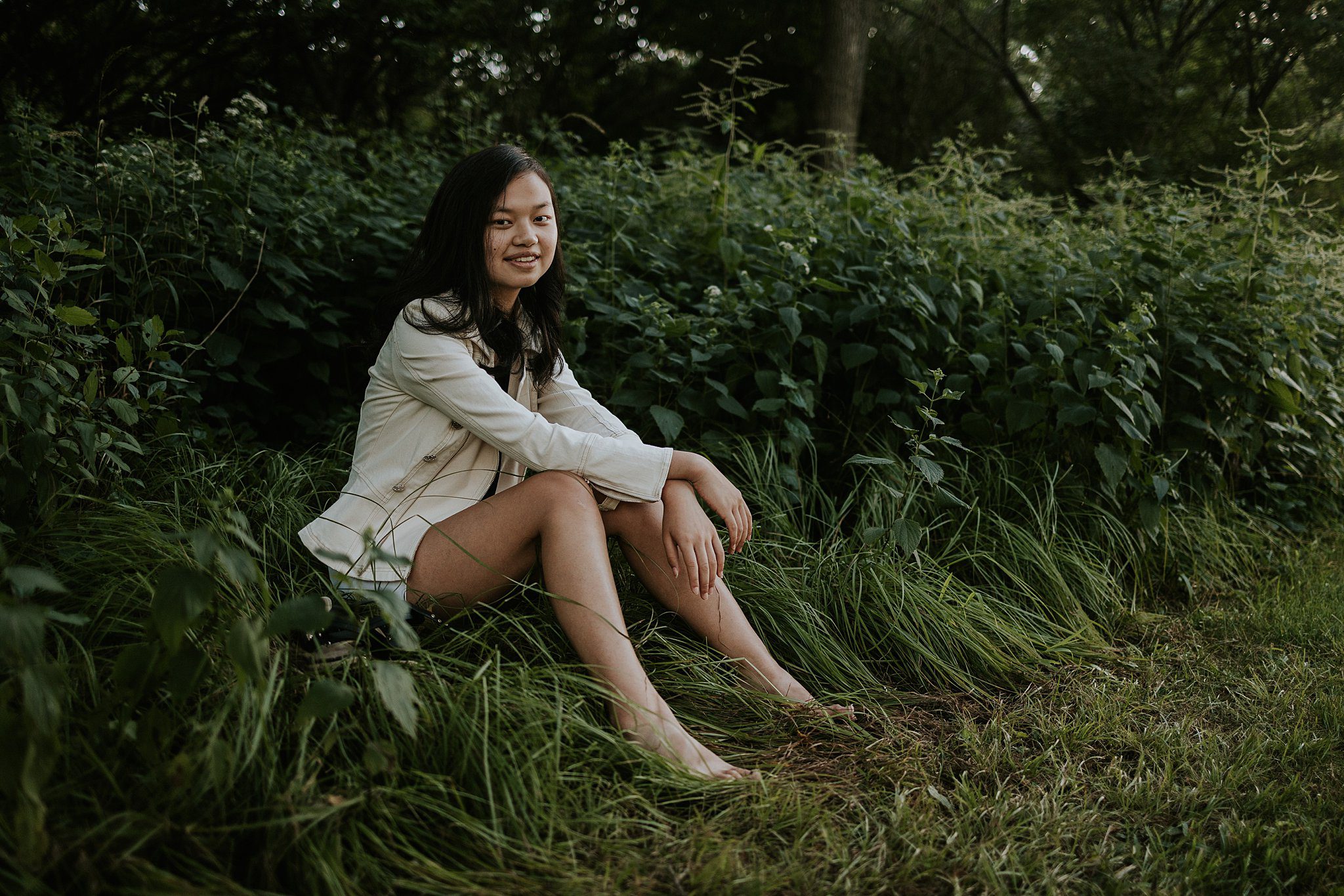 A girl is getting her senior photos taken in Northwest Iowa. She has black hair and is wearing a white jacket and shorts. She's sitting comfortably in the grass in front of some native grasses smiling at the camera. 