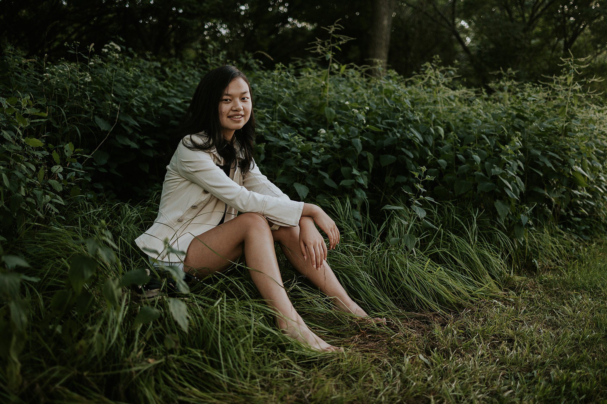 2022-08-09_0012.jpg A girl is getting her senior photos taken in Northwest Iowa. She has black hair and is wearing a white jacket and shorts. She's sitting comfortably in the grass in front of some native grasses smiling at the camera.