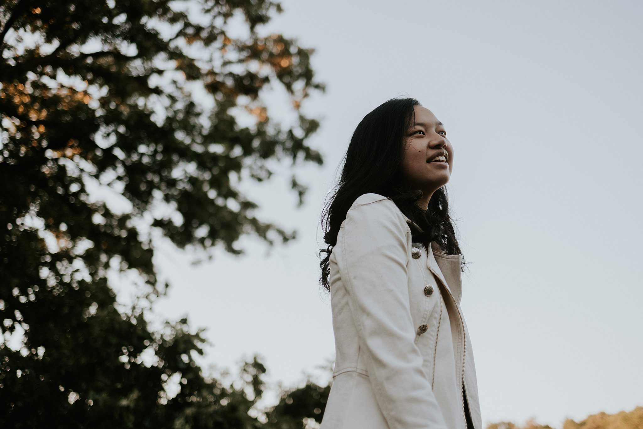 This is a photo of a girl with black hair and a white jacket taken from the ground looking up to the sky. She is smiling and looking off in the distance with a tree on the back left. 