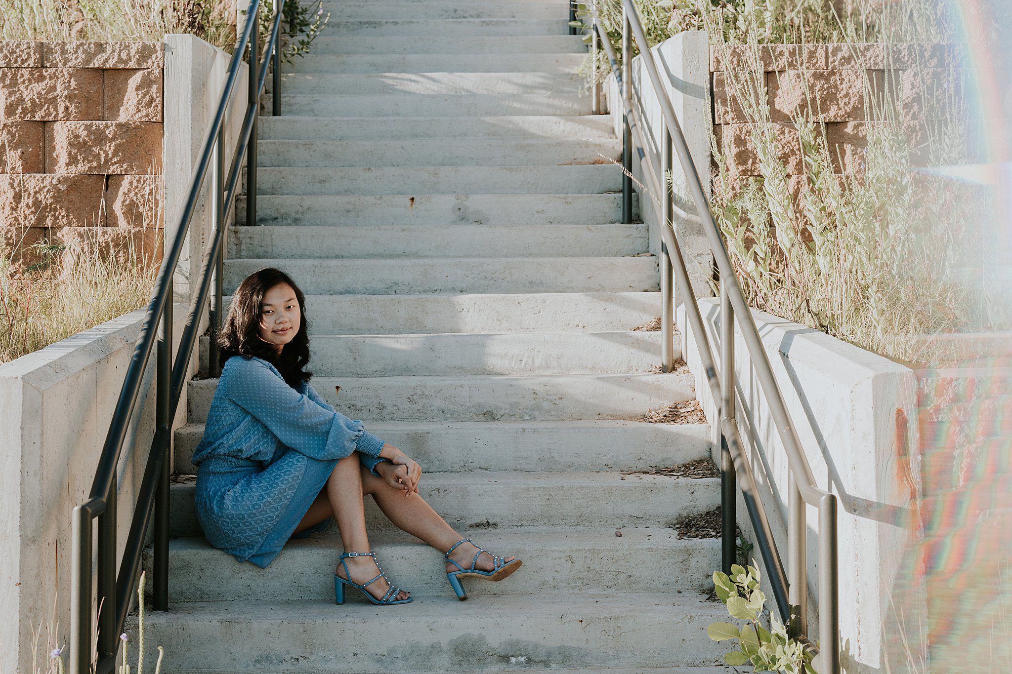 A girl with black hair in a blue dress  and blue heels is sitting on some cement stairs. On either side of the stairs there are native flowers meant to be a monarch sanctuary. The sunset is shining in from the left of the photo casting interesting shadows on the stairs and a rainbow flare is on the right side of the photo. Girl is smiling softly at the camera while getting her senior portraits taken. 