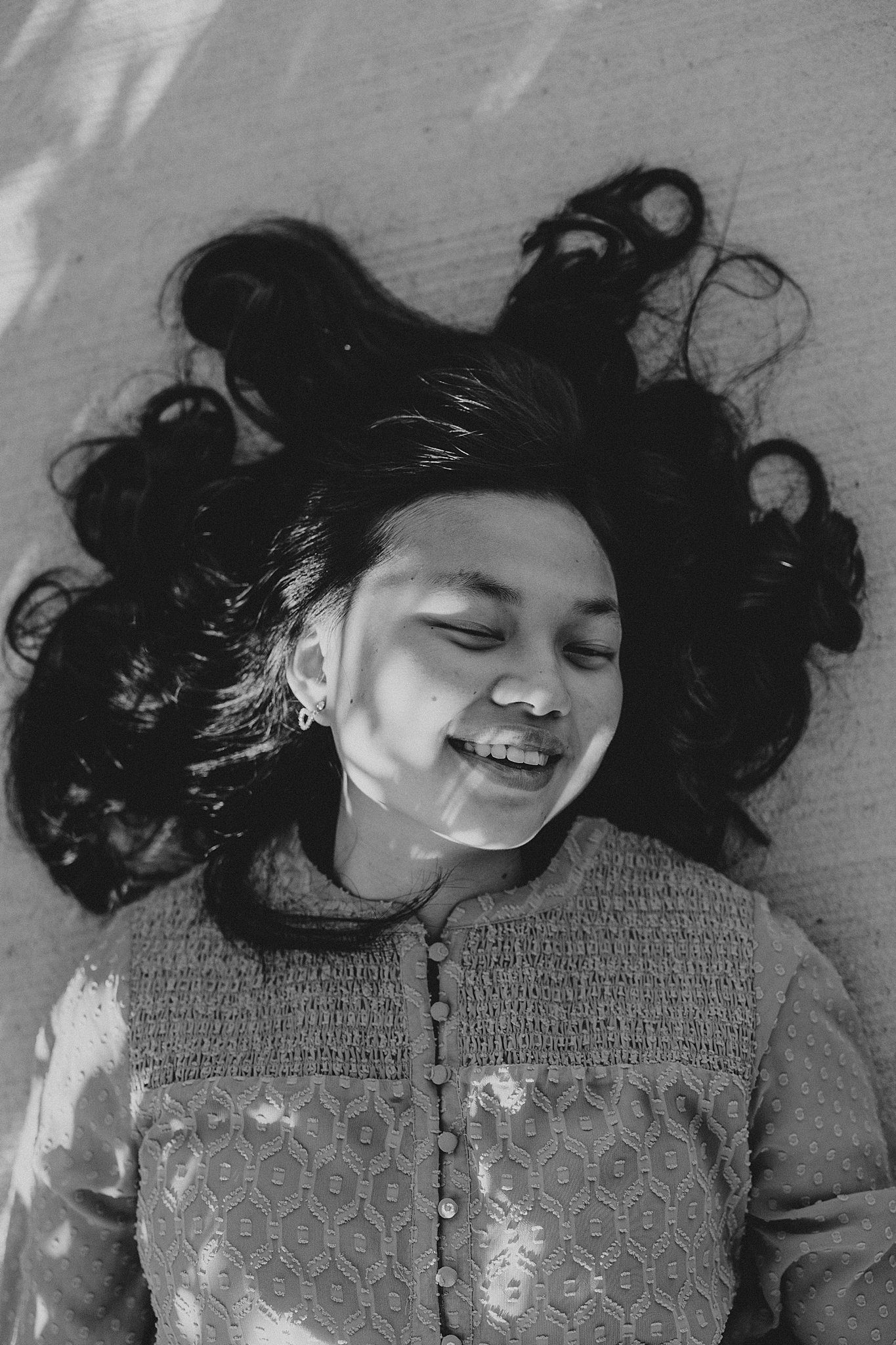 A girl with black hair laying on the ground is laughing. Light is coming through the area next to her casting interesting shadows on her face. 