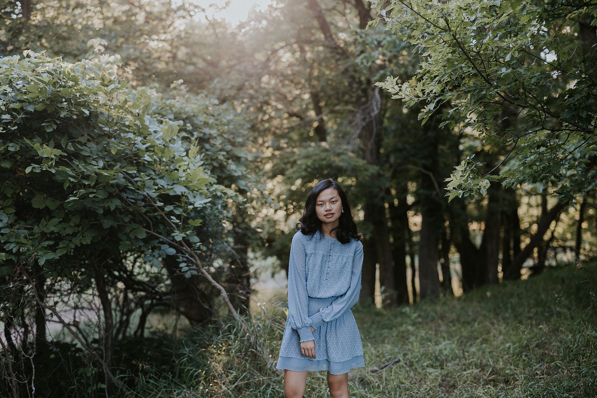 A girl with black hair is standing in the woods getting her senior portraits taken. Golden light is coming in over the trees and she is wearing a light blue dress. Senior girl looks like a model.