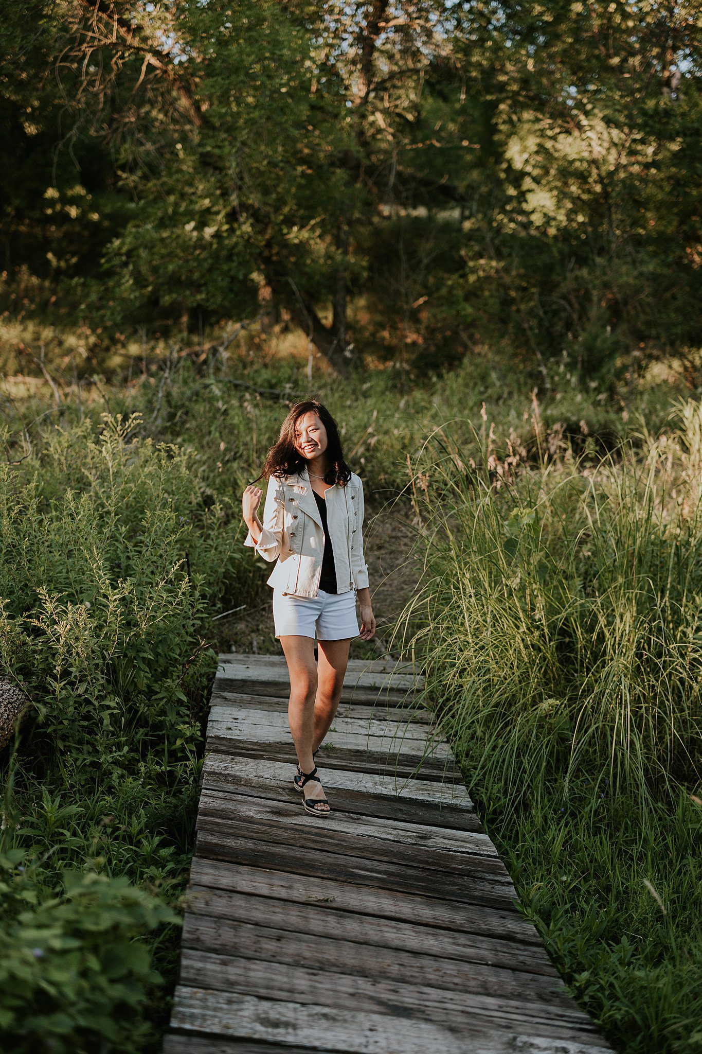 2022-08-09_0020.jpg Senior photography near Le Mars Iowa of a Girl walking across a bridge. She is smiling and playing with her hair while the sun comes in from the left and shines on her face.